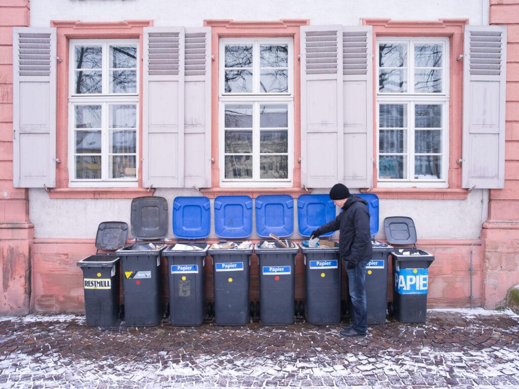 Eine Reihe von Mülltonnen vor einem Haus, organisiert und teilweise geöffnet, mit einer Person, die eine Mülltonne nutzt. Die Mülltonnenbox 2er bietet eine praktische Lösung für ein aufgeräumtes Erscheinungsbild im Garten.