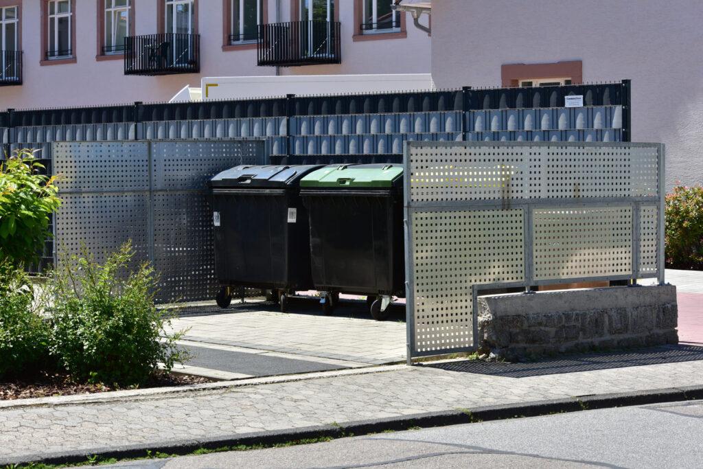 Eine Mülltonnenecke mit mehreren Mülltonnen, umgeben von einem modernen Sichtschutz. Die Mülltonnenbox 2er hilft, Ordnung und ein ansprechendes Erscheinungsbild im Garten zu bewahren.