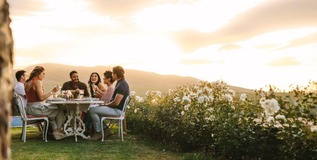 Eine Gruppe von Freunden sitzt an einem gedeckten Tisch im Garten und genießt ein Abendessen bei Sonnenuntergang, umgeben von blühenden Rosenbüschen und einer idyllischen Landschaft.