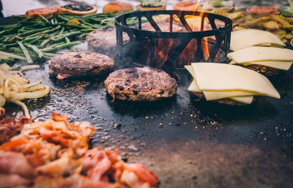 Burger-Patties und Gemüse werden auf einer heißen Feuerplatte gegrillt, während Käse schmilzt. Die Feuerplatte bietet eine vielseitige Kochmöglichkeit für Outdoor-Grillabende.