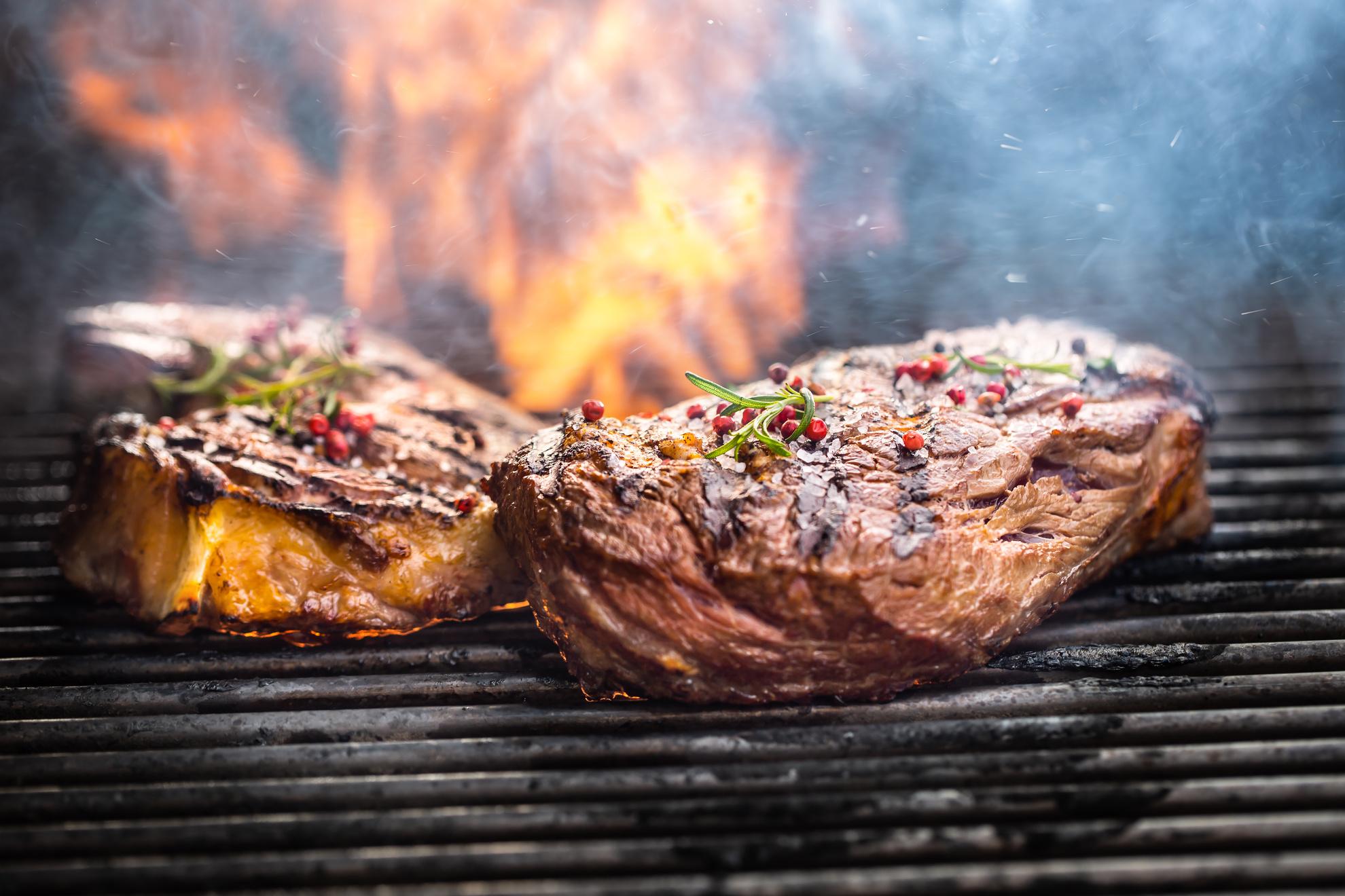 Drei saftige Steaks werden auf einem Grill zubereitet, die Flammen lodern im Hintergrund. Die Steaks sind perfekt gegrillt, mit einer knusprigen Kruste und frischen Gewürzen, darunter Rosmarin und rosa Pfefferkörner. Rauch steigt auf, während das Feuer den Grill umgibt, was eine authentische, rustikale Grillatmosphäre schafft.