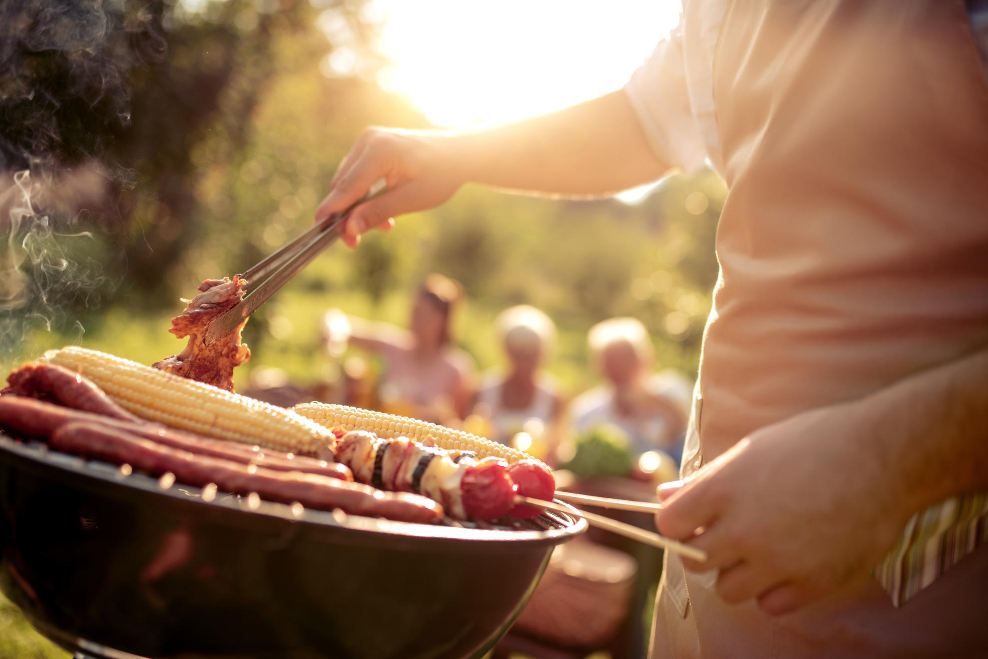 Eine Person steht am Grill und wendet Fleischstücke mit einer Zange. Auf dem Grill liegen neben dem Fleisch auch Maiskolben und Gemüse-Spieße. Im Hintergrund sitzen verschwommen mehrere Menschen bei einem sonnigen Picknick im Freien, während Rauch vom Grill aufsteigt und die goldene Abendsonne die Szene beleuchtet.