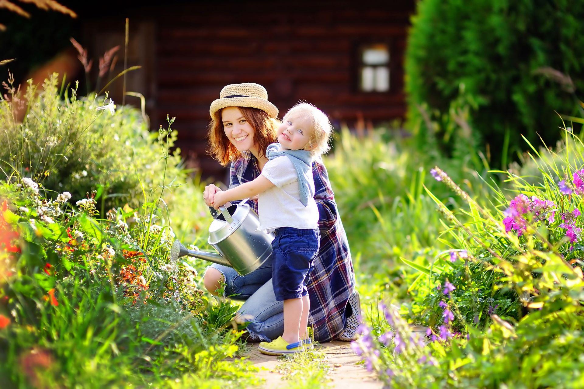 Mutter mit kleinem Kind beim gemeinsamen Gießen von Blumen in einem blühenden Garten, beide lächeln und genießen die Gartenarbeit. 