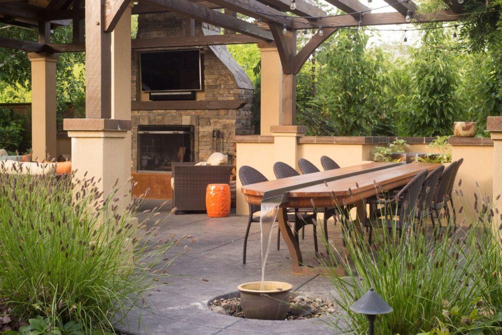 Terrassenueberdachung aus Holz mit Esstisch, Wasserlauf, Kamin und ueppigem Gruen – naturnaher Aussenbereich mit funktionalem Uebergang