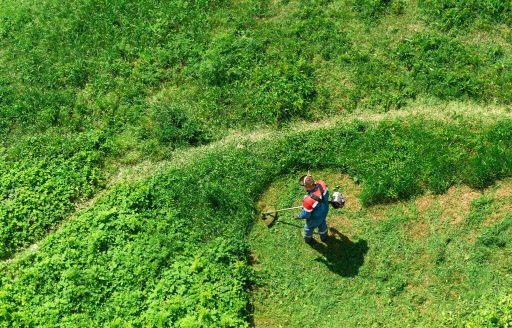 Person schneidet wild gewachsene Wiese mit einem Freischneider in unebenem Gelaende mit mehr Aufwand durch händische Arbeit